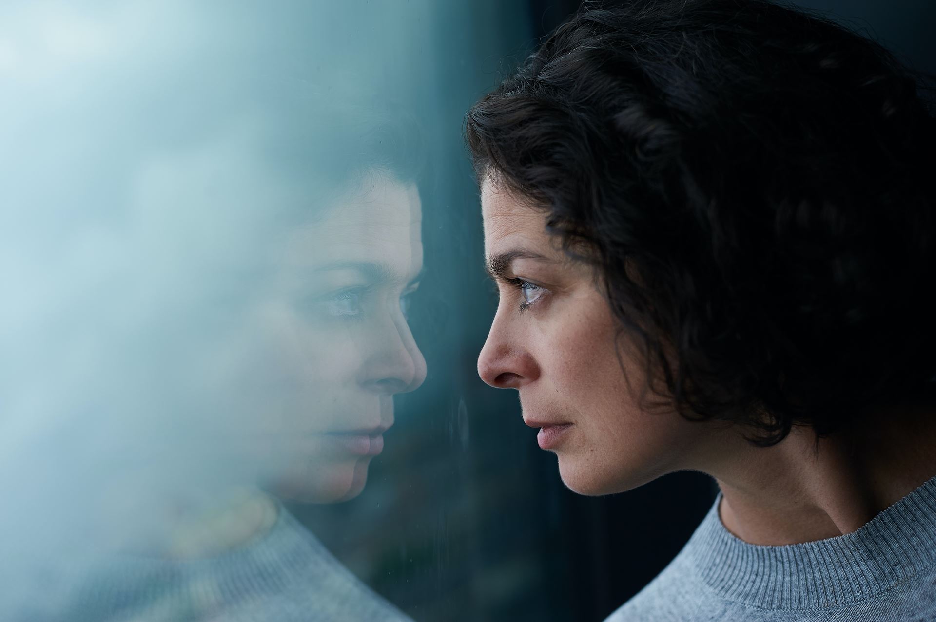 a woman looking through a window, her image reflected in the glass